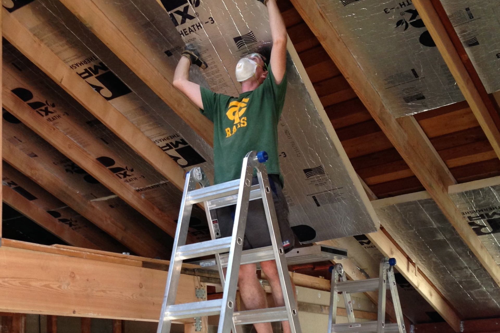 A man installs insulation on a roof, wearing safety gear and focused on his task in a clear blue sky.