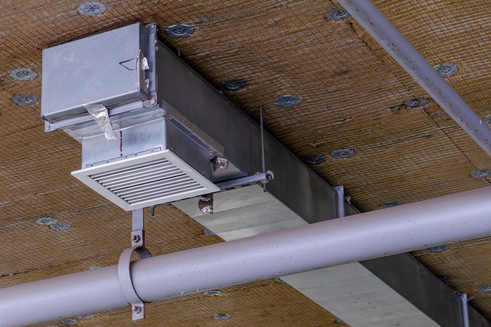 Metal ductwork and ventilation grill mounted on a ceiling, with visible pipes and textured surface above.