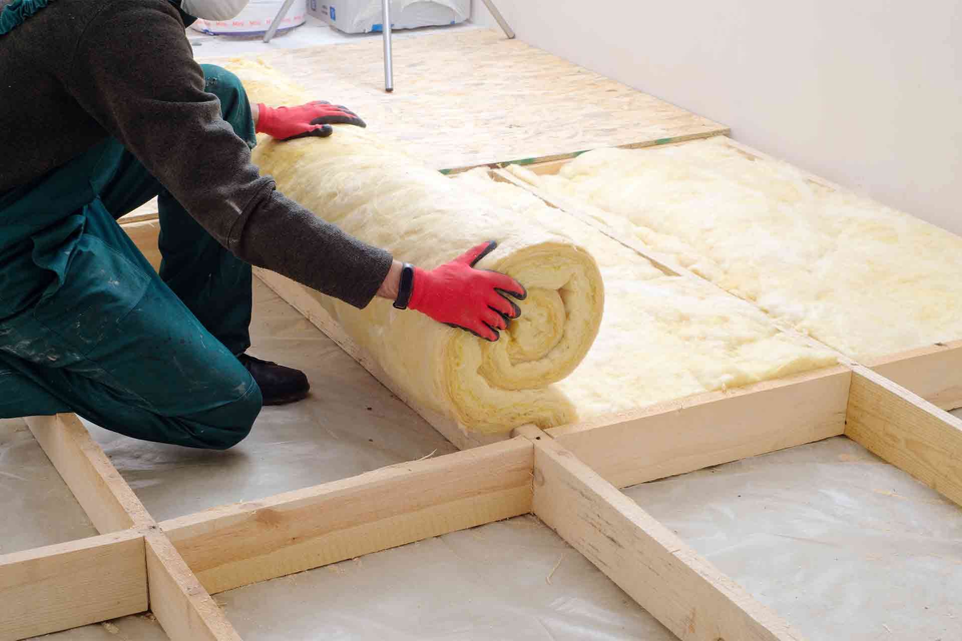 A worker in gloves rolls out insulation material onto a wooden floor framework during a home renovation project.