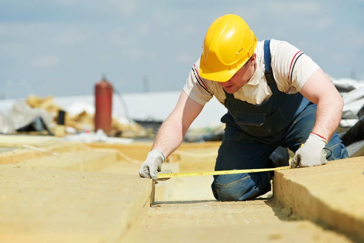A construction worker in a yellow hard hat measures a concrete slab with a tape measure on a sunny day at a building site.
