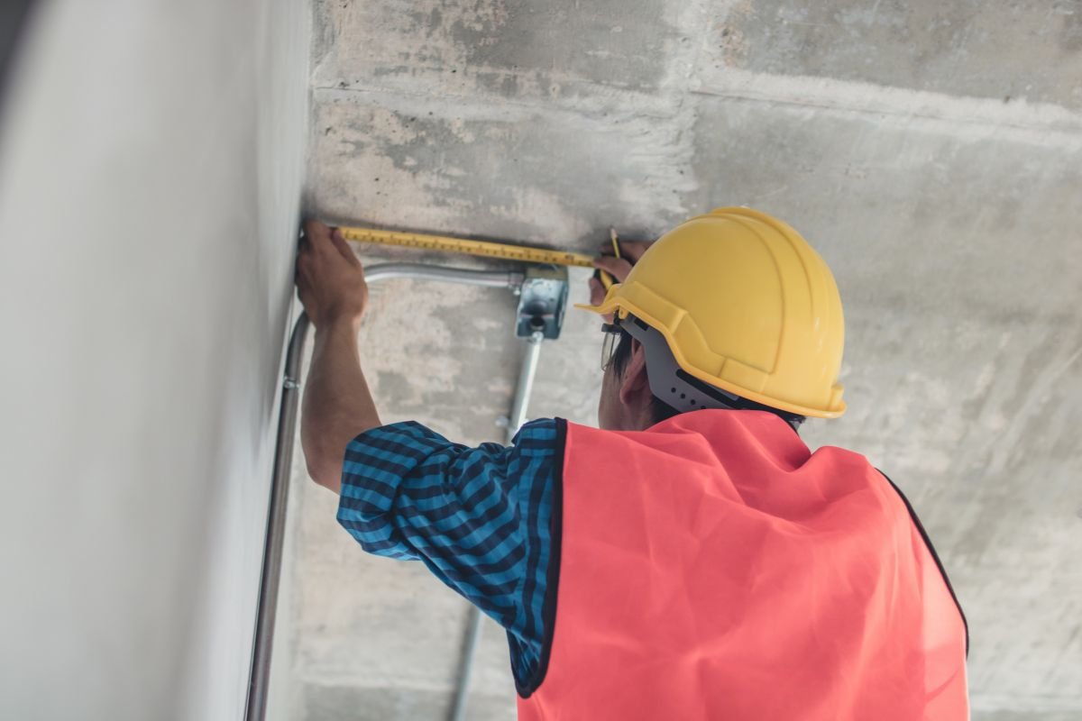 A construction worker in a yellow hard hat and orange vest measures ceiling height with a tape measure in a concrete building.