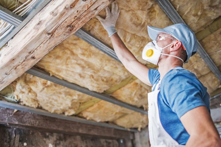 A man wearing a mask and gloves installs insulation in a ceiling while reaching above him, with exposed wood and metal beams visible.