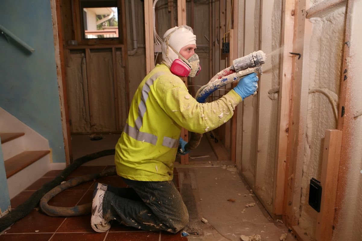 A worker in a neon yellow safety jacket kneels, using a spray nozzle to apply insulation within a partially constructed room.