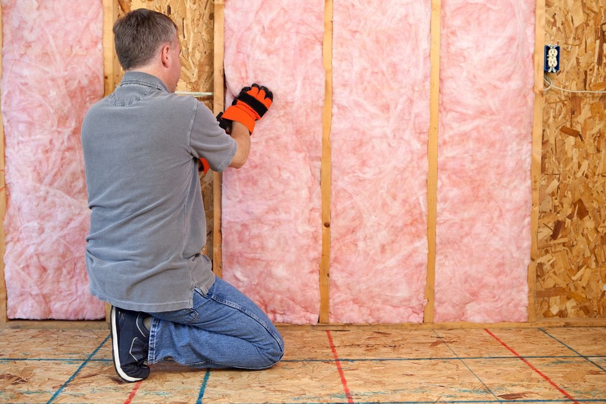 A man kneels on the floor, installing pink insulation between wooden studs in a home renovation project. He wears gloves for protection.