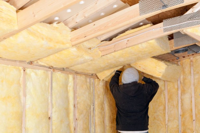 A person in a gray beanie installs yellow insulation material in the ceiling of an unfinished room with wooden framing.