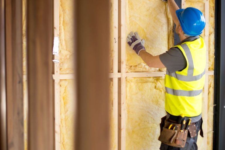 A man in a yellow vest and hard hat installs insulation on a wall, focusing on his task with tools in hand.
