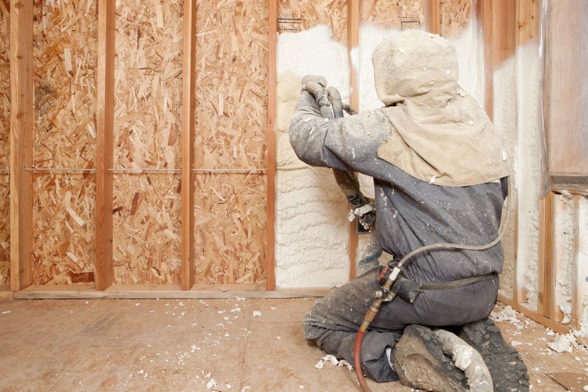 A man wearing a hard hat and jacket applies paint to a wall with fiberglass batts insulation. 